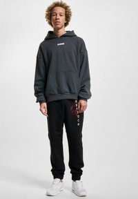 Dark gray hooded sweatshirt with front pocket, paired with black sweatpants featuring red graphic and white text, worn with white sneakers.