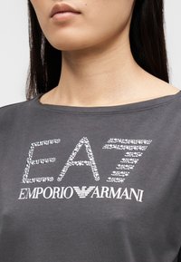 Szara bawełniana koszulka z aplikacjami z kryształków układającymi się w napisy "EA7" oraz "EMPORIO ARMANI", o luźnym kroju i szerokim dekolcie.