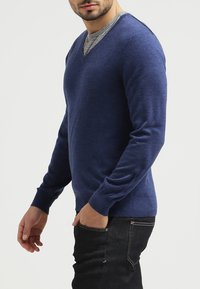 Marineblauer Pullover aus weichem Material, mit V-Ausschnitt und langen Ärmeln, kombiniert mit dunklen Jeans.