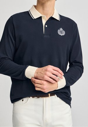 Mężczyzna ubrany w granatową koszulkę polo z długim rękawem, kremowym kołnierzykiem i mankietami, z haftowanym herbem na piersi, oraz białe spodnie z brązowym paskiem.