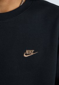 Čierna mikina so zaobleným výstrihom. Na ľavej strane hrude je malé, zlaté vyšívané logo Nike. Mäkká textúra látky.
