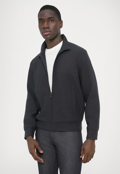 Vista frontale di una giacca nera con cerniera, motivo a trama, collo alto e polsini a coste, indossata sopra una t-shirt bianca e jeans scuri.