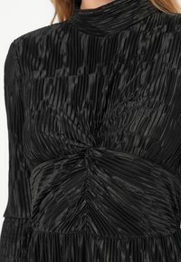 Fekete ruha pliszírozott dizájnnal, magas nyakkivágással és csavart csomó részlettel a deréknál. Simító, fényes anyagból készült, függőleges textúrával.