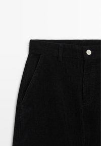 Pantalon en velours côtelé noir avec passants de ceinture, poche latérale et fermeture par bouton argenté à la taille, vu de la partie haute avant.