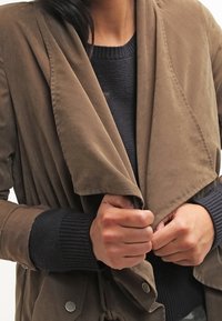 Brązowa zamszowa kurtka z opadającym kołnierzem i przednimi kieszeniami. Mankiety mają teksturowany dzianinowy wykończenie, co dodaje kontrastu gładkiej tkaninie.