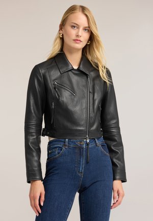 Ung kvinde med langt blondt hår iført gyldne hoops-øreringe, en sort læderjakke og blå højtaljede jeans, stående mod en neutral baggrund.