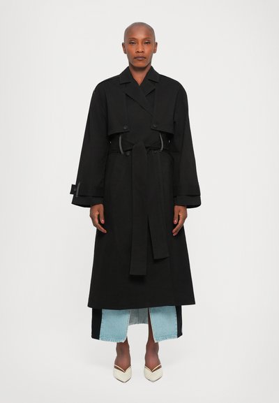 Schwarzer Trenchcoat mit strukturierter Silhouette, doppelreihigen Knöpfen und Taillengürtel. Kombiniert mit einem Jeansrock und spitzen Schuhen.