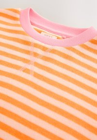 Gros plan d'un pull rayé orange et rose avec un col côtelé rose et un détail de croix cousu sous l'encolure.