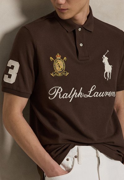 Άνδρας που φορά καφέ polo Ralph Lauren με λευκό αριθμό 3 στο μανίκι, λογότυπο της μάρκας, έμβλημα και λευκό παντελόνι με χέρι στην τσέπη.
