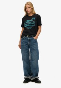 Sort bomulds T-shirt med turkis grafisk tekst; løse blå denim jeans; opsmøgede ærmer; sorte sko; neutral baggrund.