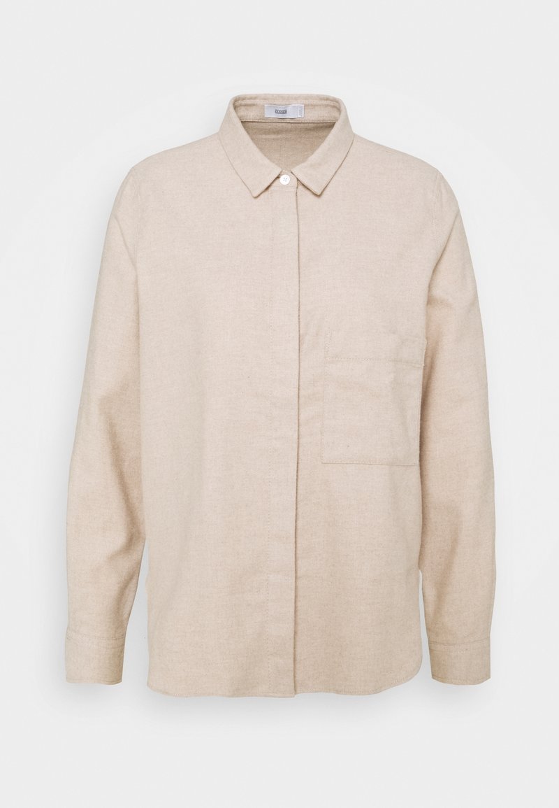 Chemise beige à manches longues avec col classique, une poche poitrine simple, patte de boutonnage cachée et poignets boutonnés sur fond blanc.