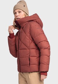 Gepolsterte, rostfarbene Jacke mit hohem Kragen und Kapuze. Verfügt über ein gestepptes Design und schwarze Reißverschlußtaschen. Getragen wird eine gestrickte beigen Mütze.