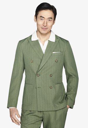 Hombre con un traje cruzado de rayas verdes y una camisa blanca con cuello, de pie con una mano en el bolsillo sobre un fondo blanco.