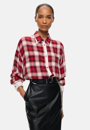 Mujer que lleva una camisa de cuadros roja y blanca abotonada metida en una falda de cuero negra con un cinturón negro, de pie con una mano en el bolsillo.