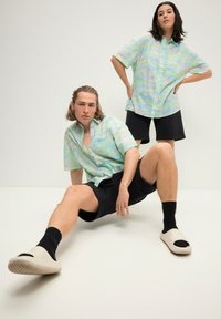 Kortærmede, knap-op skjorter med et pastelfarvet grønt og lilla mønster, parret med sorte shorts. Model i lette slip-on sko.
