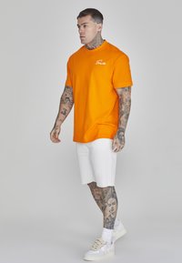 Orange bomulds t-shirt med rund hals, korte ærmer og hvid broderet logo, bæres med hvide shorts og sneakers. Tatoveringer synlige på armene.
