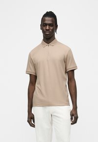 Homme aux cheveux tressés portant un polo beige côtelé à manches courtes et un pantalon blanc, debout devant un fond blanc uni.
