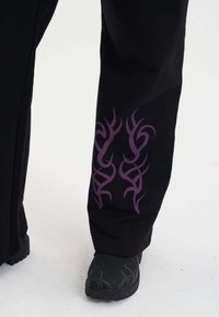 Zwarte broek met paarse tribale ontwerpen op de onderbenen. Materiaal lijkt textuur te hebben. Draagt bij donkere schoenen met een vlampatroon.