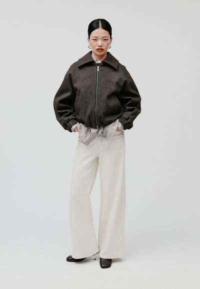 Giacca bomber marrone con zip e polsini elasticizzati, indossata su una camicia a righe. Abbinata a pantaloni larghi color crema e stivaletti neri con tacco.