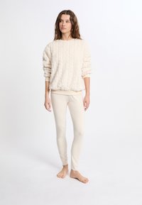 Sweter w kolorze kremowym, puszysty, z ribbowanymi mankietami, w połączeniu z jasnobeżowymi dopasowanymi legginsami. Obie części odzieży mają miękką teksturę i luźny krój.