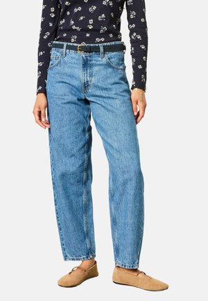 Person iført højtaljede, lige blå jeans med sort bælte, sort blomstrede langærmet top, brune flade sko og et fint armbånd på det ene håndled.
