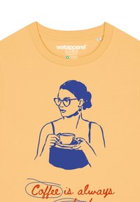 Geel katoenen t-shirt met een blauwe graphic van een vrouw die een kopje vasthoudt, met de tekst "Koffie is altijd" in het rood eronder.
