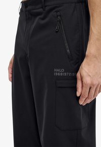 Zwarte broek gemaakt van een glad, lichtgewicht materiaal met een ritssluitingzak, decoratieve stiksels en een zijvak met logo details.