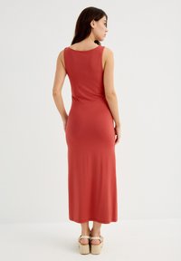Vestido largo en color óxido, hecho de tela elástica. Presenta un diseño sin mangas, con escote en la espalda y un corte entallado, combinado con sandalias de plataforma.