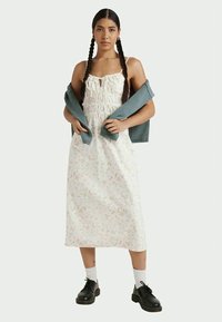 Junge Frau mit geflochtenem Haar, die ein weißes, floral gemustertes Midikleid trägt, schwarze Schuhe mit weißen Socken und einen blauen Pullover um die Arme hält.