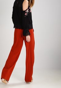 Svart off-the-shoulder blus med blommig broderi och spetsdetaljer, ihopparad med vida röda byxor. Mjuk tygkvalitet och avslappnad passform.