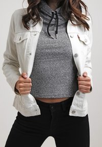 Valkoinen denim-takki harmaan lyhyen hupparin päällä, jossa on nauhakotelo, näkyy mustia farkkuja. Tekstuuri on sileä ja rento.
