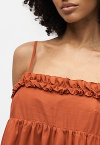 Γυναίκα που φοράει ένα αμάνικο φόρεμα σε σκουριά-πορτοκαλί χρώμα με λεπτές τιράντες και βολάν κατά μήκος του ντεκολτέ και της μέσης.