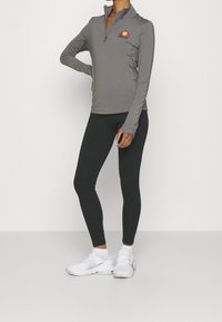 Šedý top na zip s dlouhým rukávem a logem na hrudi, kombinovaný s černými legínami a bílými sportovními botami, na jednoduchém pozadí.