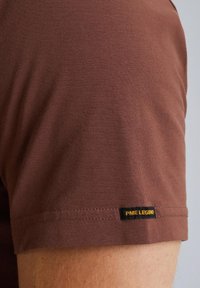Braunes Kurzarm-T-Shirt aus weichem Material, mit einem schwarzen Etikett und gelbem Text "PME LEGEND" am unteren Saum.