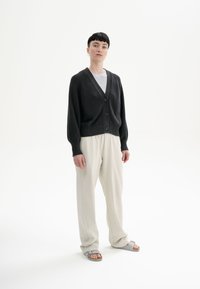 Dunkelgraue Strickjacke mit V-Ausschnitt, Knopfverschluss, gerippten Bündchen, kombiniert mit hellbeigen weiten Hosen und grauen Sandalen.