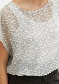 Blusa bianca con un motivo trasparente a onda in nero. Collo rotondo e maniche corte, vestibilità leggermente ampia con orlo curvo. Parte inferiore nera visibile.