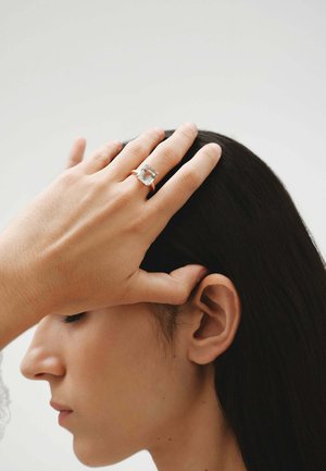 Anillo de plata con una gran piedra rectangular clara, llevado en una mano que descansa contra una cabeza. El tono de piel es claro; el pelo es oscuro y está peinado hacia atrás.