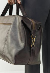 Bolso de cuero marrón oscuro con textura suave, cremallera dorada y dos asas robustas. Cuenta con una correa desmontable con gancho de presión.