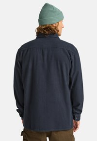 Marineblauw, lange mouwen blouse met een klassieke kraag, knopenmanchetten en een rechte zoom; gemaakt van structuurstof, gedragen met een groene been.