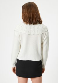 Weißer gestrickter Pullover mit einer strukturierten, gepunkteten Passe und gekräuseltem Rand; über einem schwarzen Rock getragen, was kontrastierende Farben und Materialien zur Geltung bringt.