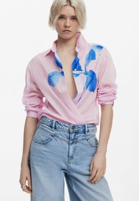 Camicia button-up rosa chiaro con design floreale blu, maniche arrotolate, indossata con jeans azzurri a vita alta, caratterizzati da una vestibilità comoda.