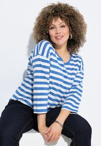 Gestreiftes blau-weißes Langarmshirt mit Rundhalsausschnitt, kombiniert mit dunklen Jeans. Das Model hat lockige Haare und sitzt.