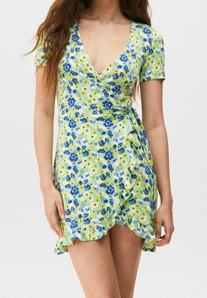 Mujer con un vestido corto cruzado con estampado floral de flores azules y verdes y dobladillo con volantes, de pie frente a un fondo liso.