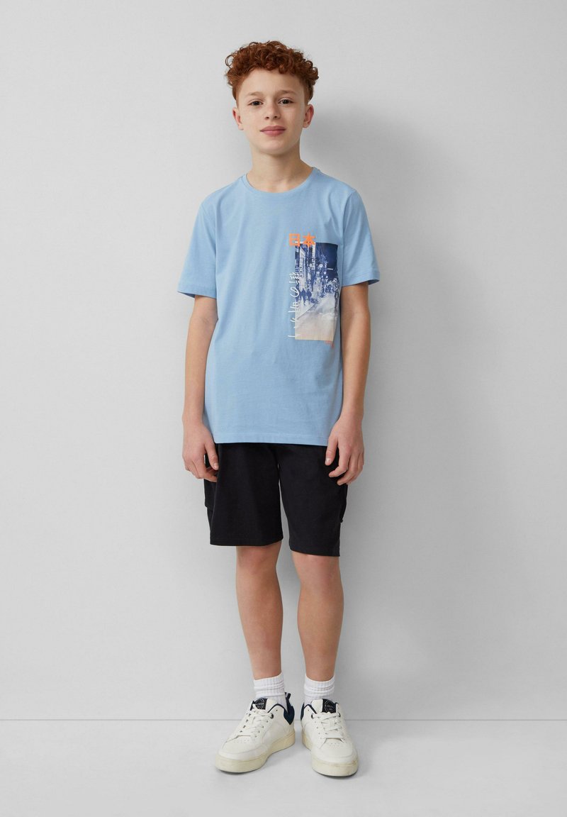 Lichtblauwe katoenen T-shirt met een grafische print in blauw, oranje en wit. Gecombineerd met zwarte shorts en witte sneakers.