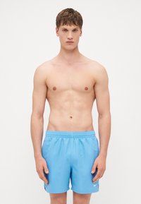 Shorts de bain bleus avec une taille élastique et un petit logo blanc sur le côté, texture lisse et longueur au-dessus du genou.