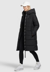 Lang zwarte pufferjas met een capuchon en witte logo-accenten langs de rits. Gemaakt in combinatie met zwarte leggings en witte high-top sneakers.