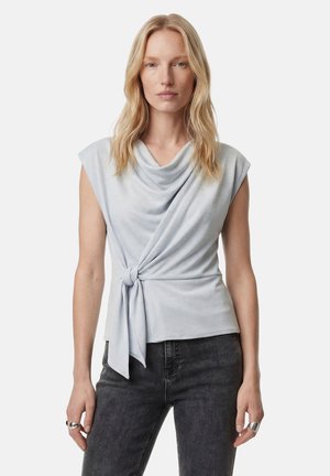 Lyshåret kvinde iført en ærmeløs, lysegrå top med faldet halsudskæring og knude foran i siden, kombineret med mørkegrå jeans.