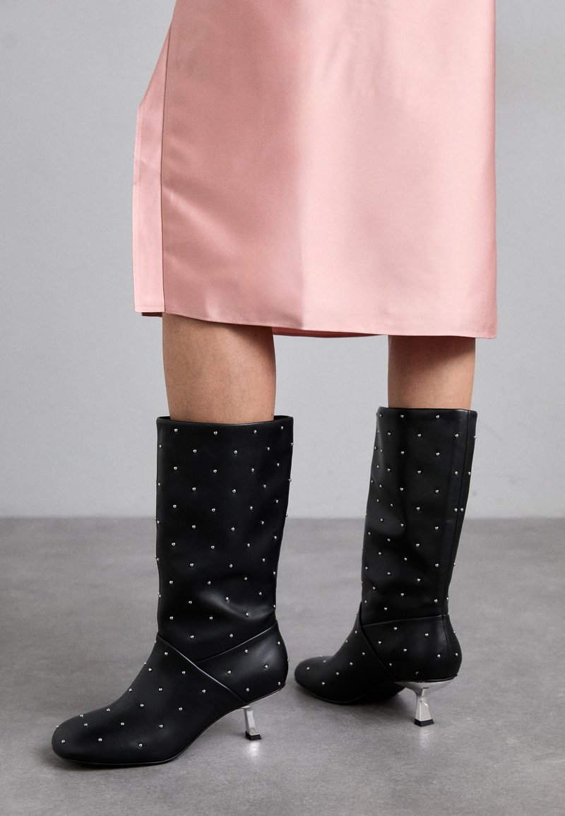 Črne škornje do kolen, izdelane iz gladkega materiala, z diskretnim križnim vzorcem in srebrnim peto. V kombinaciji z rožnato satenasto obleko.