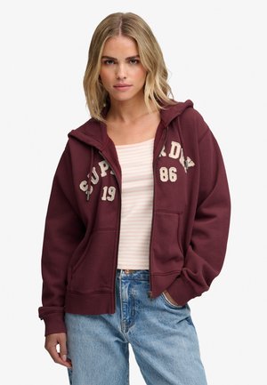 Ung kvinde med blond hår iført en vinrød hættetrøje med hvid tekst, en lys stribet top og blå jeans, stående med den ene hånd i lommen.