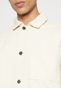 Beżowa kurtka wykonana z tkaniny o fakturze, z kołnierzem, dwoma guzikami i kieszenią na piersi. Elementy ozdobne w odcieniu srebra.